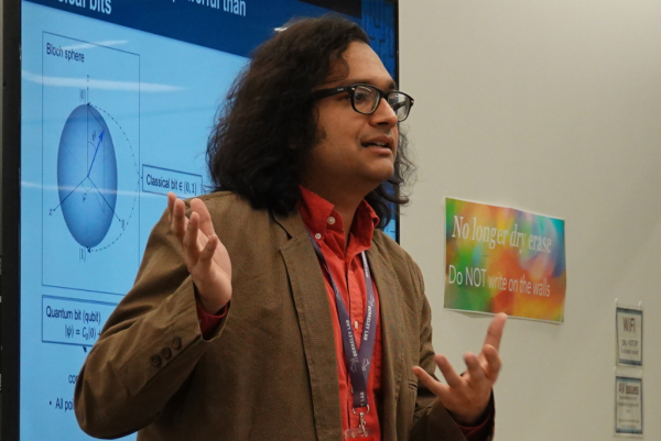 A person with shoulder-length curly hair, wearing glasses, a red shirt, and a brown blazer, is giving a presentation. They are gesturing with both hands while speaking. Behind them, a large screen displays a diagram of the Bloch sphere, which is used in quantum computing to represent qubit states.