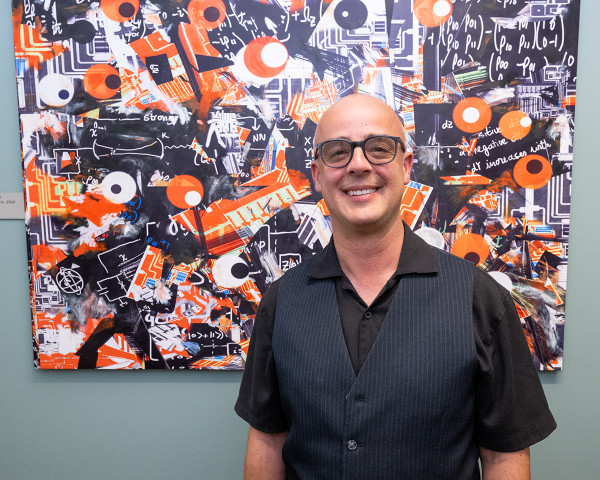 Benjamin Arizmendi in a black shirt and vest in front of a large painting containing black, orange, and white shapes.