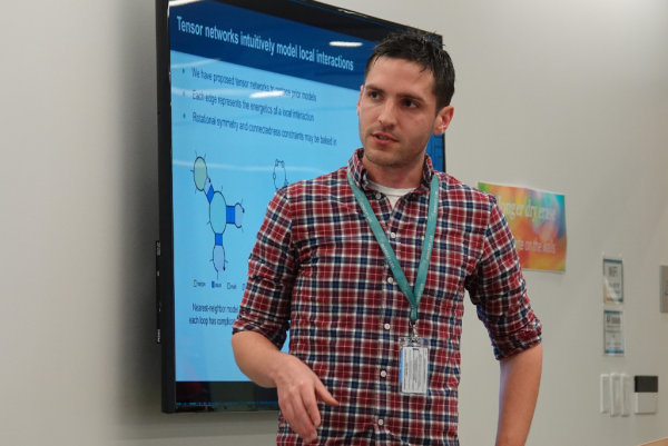 A man with short dark hair, wearing a red plaid shirt and a lanyard with an ID badge, is giving a presentation. He is gesturing with his right hand while speaking. Behind him, a large screen displays a slide about tensor networks, featuring diagrams of network structures and text explaining local interactions.