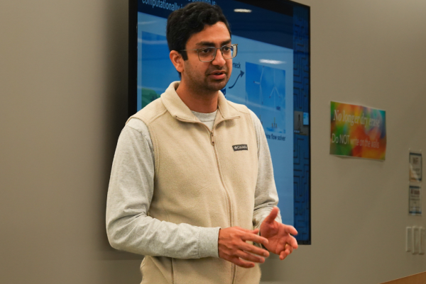 A person wearing a light-colored fleece jacket and a gray long-sleeve shirt is standing and gesturing with their hands. Behind them, there is a large screen displaying a presentation with blue graphics and text.