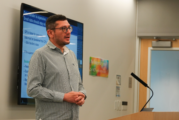 A person wearing a striped button-up shirt is standing in front of a large screen displaying a presentation slide about data transfer, GPU computation, and solutions for supercomputing. The person is positioned near a podium with a microphone. A colorful sign with partially readable text is visible on the wall to the right, and a doorway is seen in the background.