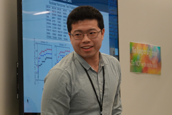 A person wearing a light gray button-up shirt and a black lanyard stands in front of a large screen displaying graphs and data.