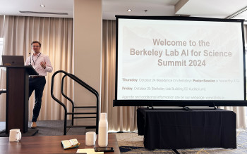 Person standing on stage next to a screen with the words Welcome to the Berkeley Lab AI for Science Summit 2024 projected onto it.