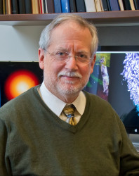 man with grey hair in a green sweater, collared shirt, and tie sitting in front of a combustion simulation 